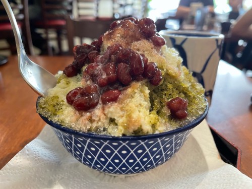A bowl of shaved ice topped with sweet red beans and a green syrup, served on a napkin in a cozy café setting.