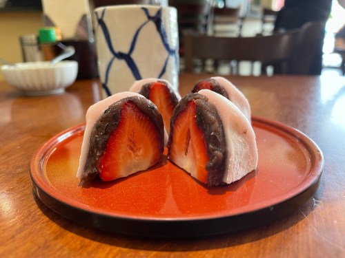 Four pieces of mochi filled with red bean paste and fresh strawberries, served on a red plate with a cup in the background.