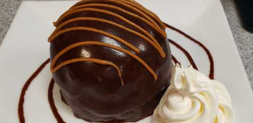 A round chocolate dessert drizzled with caramel, served with a swirl of whipped cream on a white plate.