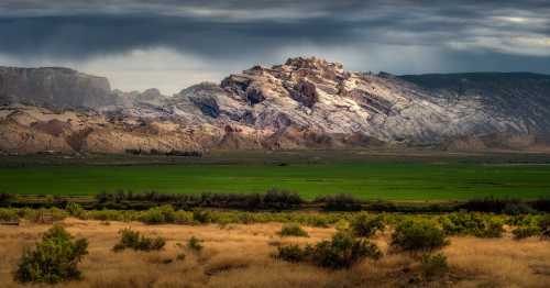 A dramatic mountain range under cloudy skies, with green fields and dry grass in the foreground.