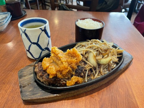 A sizzling plate with grilled meat topped with orange sauce, stir-fried noodles, and a bowl of rice, alongside a cup of tea.