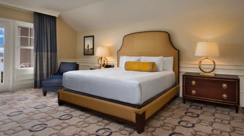 A cozy hotel room featuring a large bed, nightstands, a chair, and large windows with natural light.
