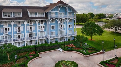 A blue hotel building with balconies, surrounded by lush gardens and a pathway, under a partly cloudy sky.