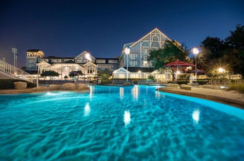 A serene pool at night, illuminated by lights, with a charming building in the background surrounded by trees.