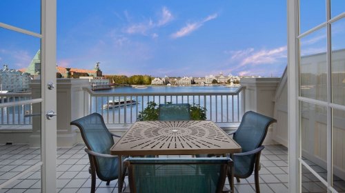 A balcony with a table and chairs overlooks a scenic waterfront view with buildings and a clear blue sky.