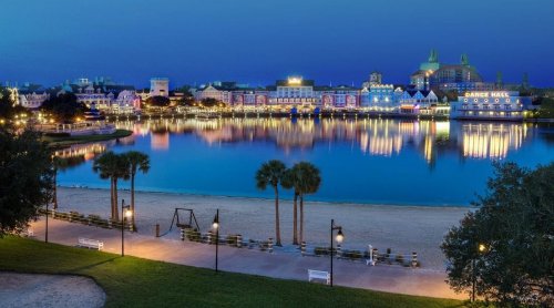 A serene lakeside view at dusk, featuring colorful buildings and palm trees reflecting on the water.