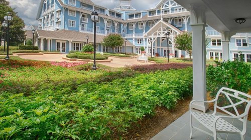 A charming blue hotel with landscaped gardens and a white chair in the foreground, under a cloudy sky.