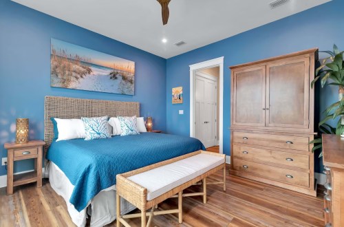 A cozy bedroom with blue walls, a large bed, wooden furniture, and a beach-themed artwork above the bed.