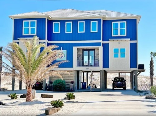 A large blue beach house on stilts, surrounded by palm trees and a driveway with a parked car.