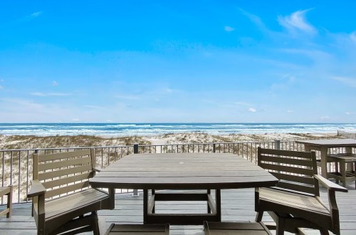 A wooden table and chairs on a deck overlooking a sandy beach and ocean under a clear blue sky.