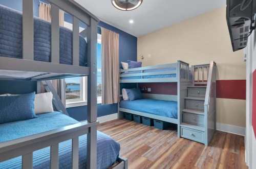 A cozy bedroom featuring a bunk bed and a twin bed, with blue bedding and wooden floors, illuminated by natural light.