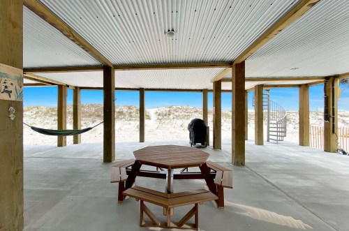 A spacious outdoor area with a wooden picnic table, hammock, and spiral staircase, overlooking sandy dunes and blue sky.