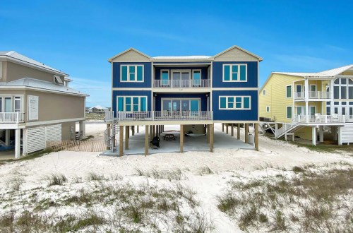 A large blue beach house on stilts, surrounded by sandy ground and other colorful homes under a clear blue sky.