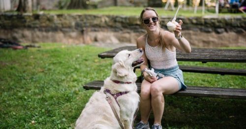 A woman sits on a picnic table with a golden retriever, holding ice cream and smiling in a grassy park.