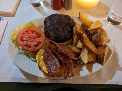 A plate with a burger topped with bacon and cheese, lettuce, tomato, and a side of crispy potato wedges.