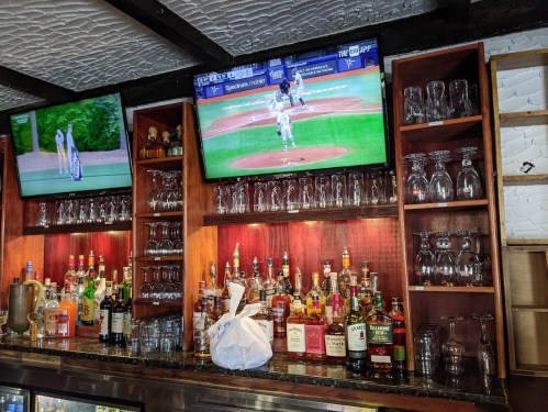 A bar shelf filled with various liquor bottles and glasses, with two TVs showing sports games in the background.