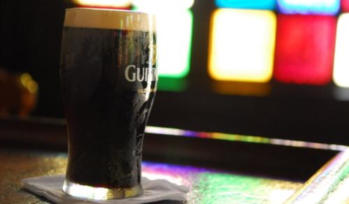 A pint of dark beer with a creamy head sits on a bar, illuminated by colorful stained glass in the background.