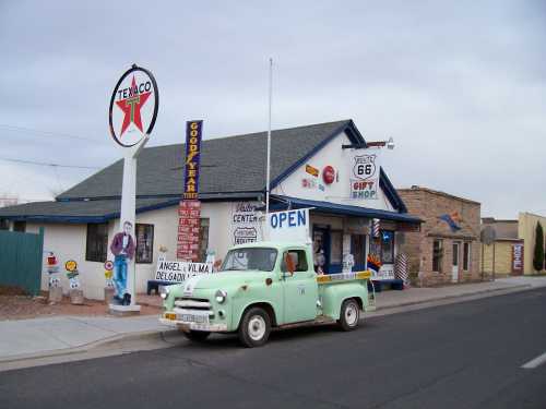 A vintage green truck parked in front of a retro gas station and gift shop with Route 66 signage.