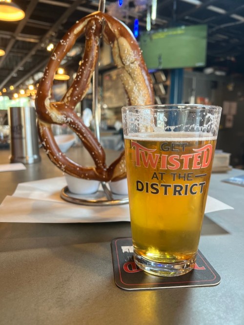 A glass of beer sits on a table next to a large pretzel on a stand in a lively bar setting.