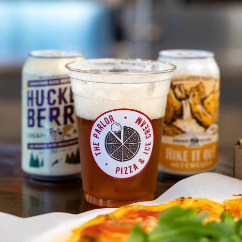 A clear cup of beer sits in front of pizza, with two colorful cans of craft beer in the background.