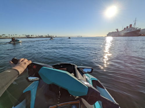 A person rides a jet ski on calm water, with a ship and palm trees in the background under a bright sun.