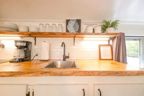 A modern kitchen with a wooden countertop, sink, coffee maker, and open shelves displaying dishes and plants.