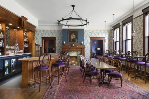 Elegant bar interior with wooden furnishings, purple seating, a fireplace, and patterned wallpaper. Cozy atmosphere.
