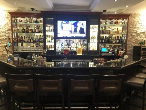 A stylish bar with a variety of liquor bottles, a large screen, and seating along the counter.