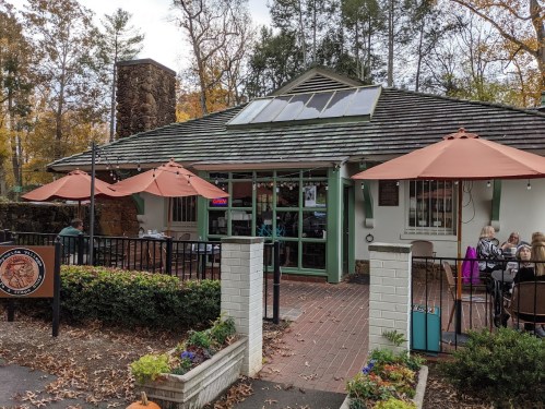 A cozy restaurant with a stone chimney, outdoor seating under orange umbrellas, surrounded by autumn foliage.