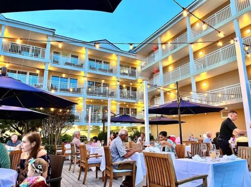 Outdoor dining scene at a hotel with guests enjoying meals under string lights and umbrellas in a cozy atmosphere.