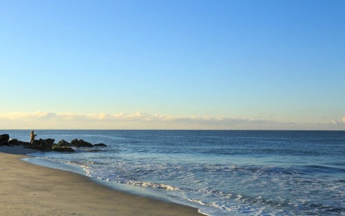 A serene beach scene with gentle waves, a clear blue sky, and a lone figure fishing near the rocky shore.