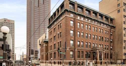 Historic brick building at street corner, surrounded by modern skyscrapers in an urban setting.