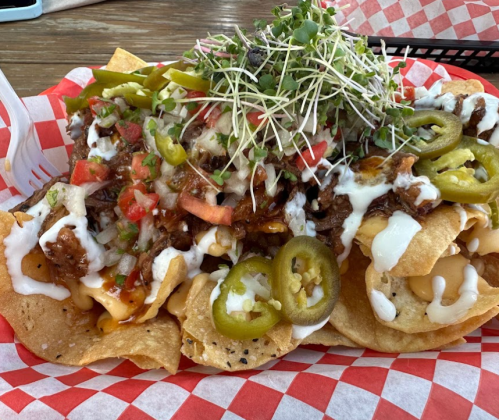A plate of nachos topped with cheese, meat, jalapeños, tomatoes, sour cream, and sprouts on a red checkered paper.