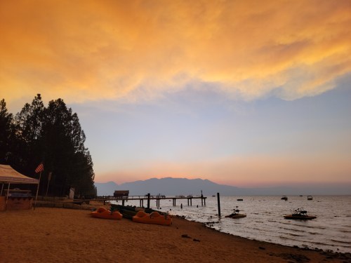 A serene beach at sunset with orange skies, a wooden pier, and boats on the water, surrounded by trees.