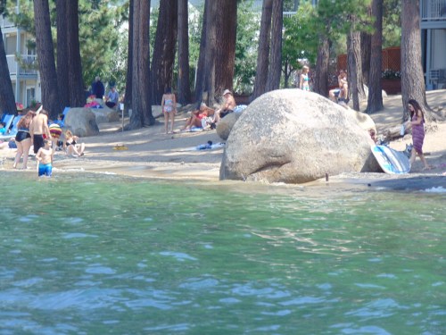 A beach scene with people sunbathing, swimming, and relaxing near large rocks and trees by the water.