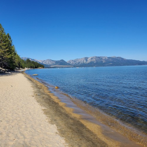 A serene beach scene with sandy shore, clear blue water, and mountains in the background under a bright blue sky.