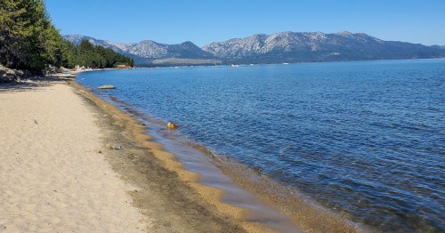 A sandy beach along a calm lake, with mountains in the background under a clear blue sky.