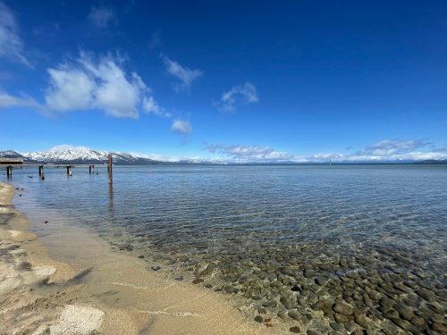 A serene lakeside view with clear water, sandy shore, and snow-capped mountains under a blue sky with scattered clouds.