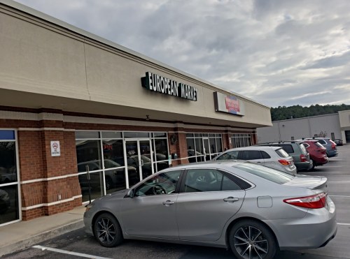 Exterior of a European market with a silver car parked in front, cloudy sky above, and several cars in the lot.
