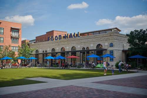 A vibrant food hall with outdoor seating, surrounded by greenery and colorful umbrellas, under a clear blue sky.