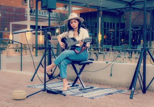 A woman plays guitar on a stage outdoors, wearing a hat and casual attire, with a microphone and speakers nearby.