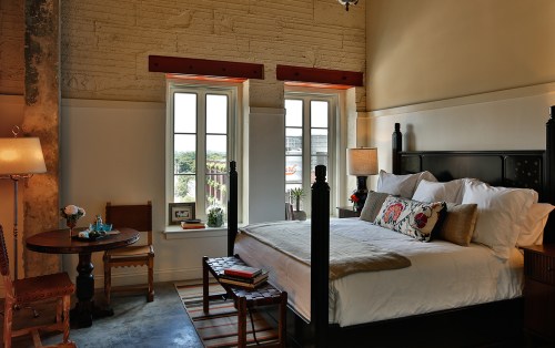 Cozy bedroom with a four-poster bed, wooden accents, and large windows, featuring a small dining table and decorative plants.