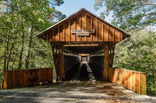 A wooden covered bridge surrounded by trees, with a sign reading "Swann Bridge" at the entrance.