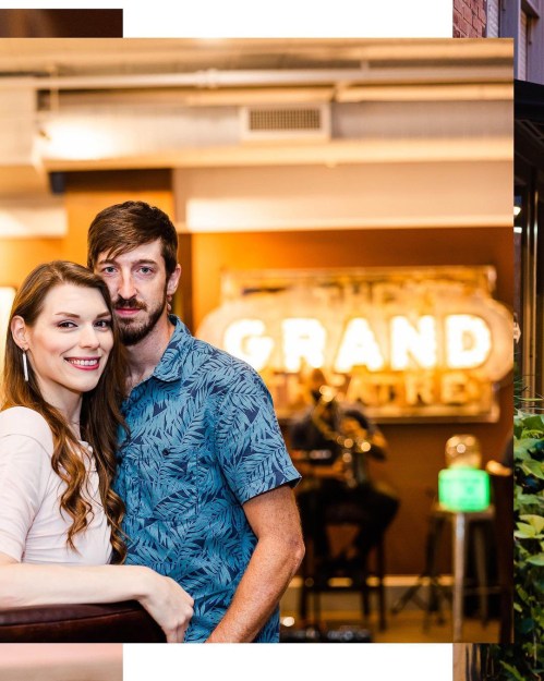 A couple poses together in a cozy venue with a "Grand" neon sign in the background, enjoying a lively atmosphere.