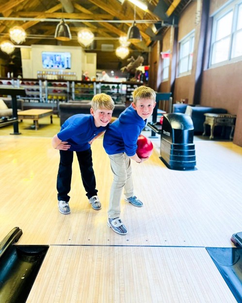 Two boys in blue shirts pose playfully at a bowling alley, one holding a red bowling ball, ready to bowl.