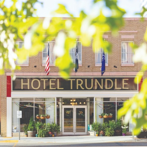 A charming hotel facade with "Hotel Trundle" sign, surrounded by greenery and flags, in a sunny urban setting.