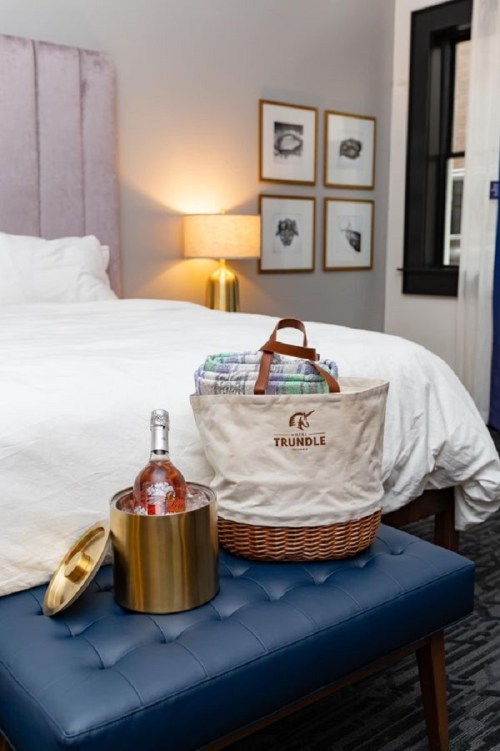 A cozy bedroom scene featuring a bed, a basket with blankets, and a bottle of sparkling wine on a blue ottoman.