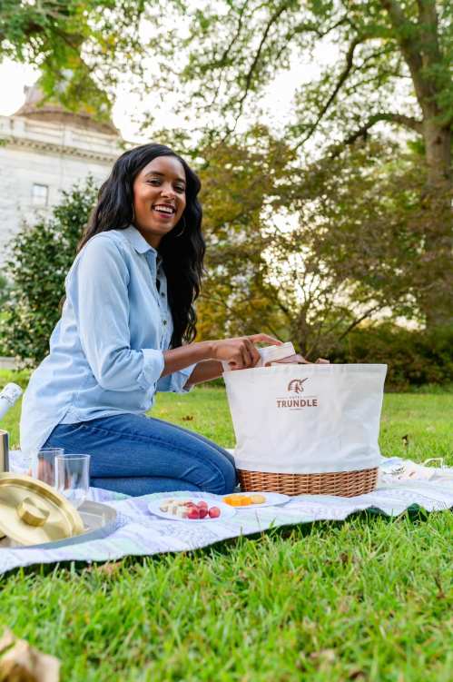 A woman smiles while unpacking a picnic basket on a blanket in a grassy area, surrounded by food and drinks.