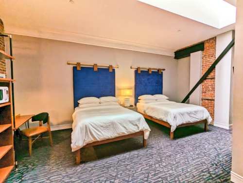 A cozy hotel room featuring two beds, a desk, and exposed brick walls, with natural light from a skylight.