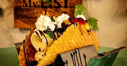 A waffle taco filled with rolled ice cream, topped with whipped cream, chocolate drizzle, and colorful fruit.